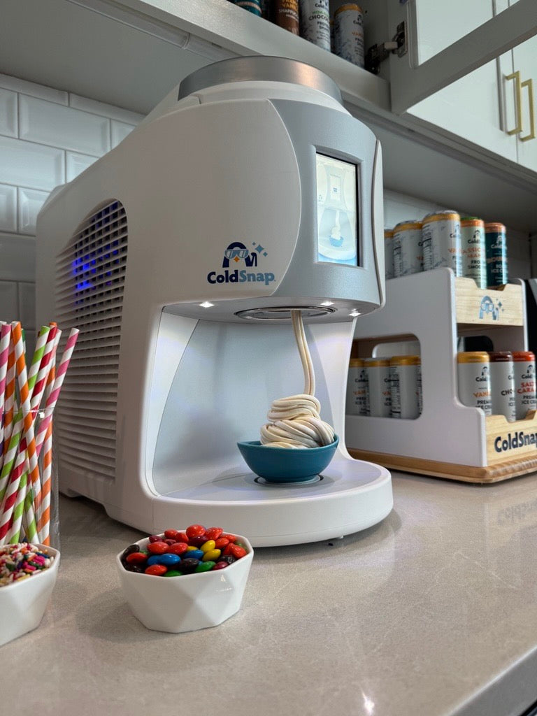 ColdSnap® frozen treat machine sitting on a kitchenette counter next to a ColdSnap® pod display rack and toppings