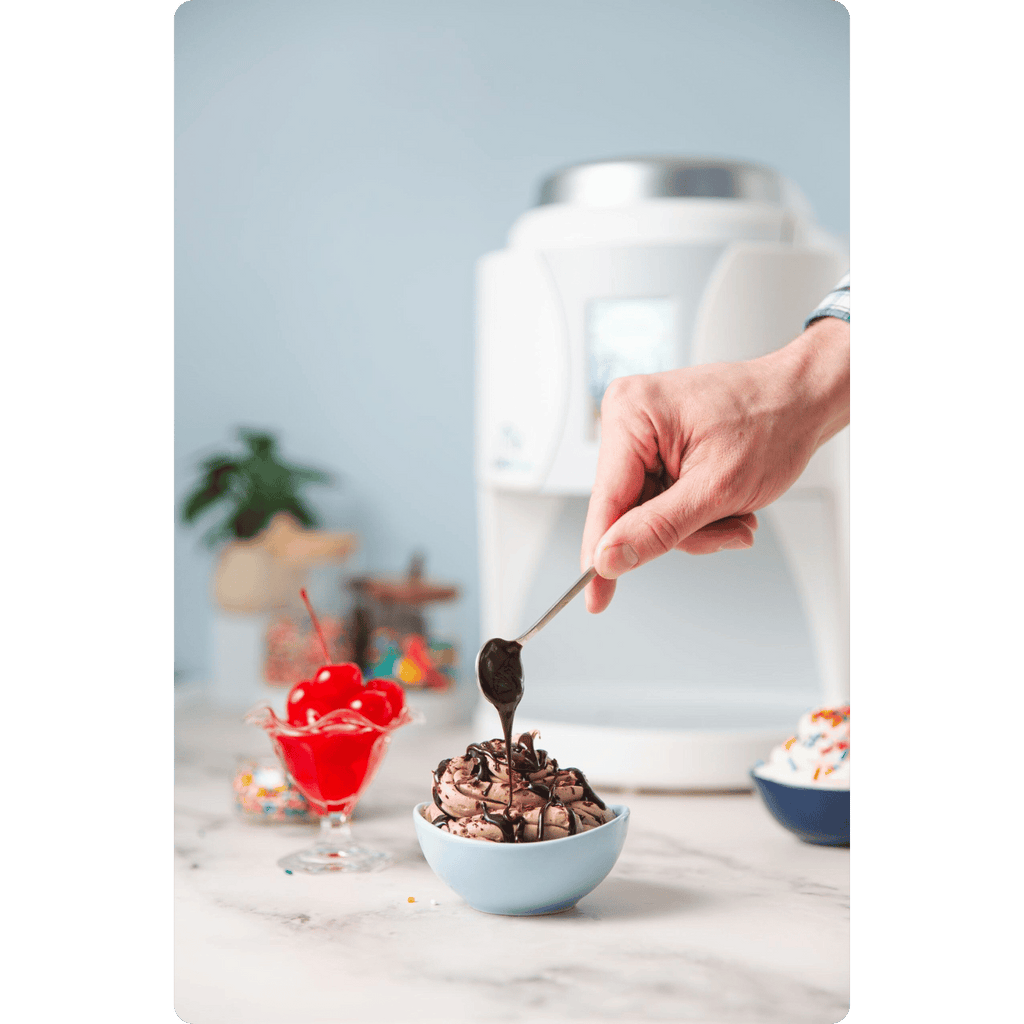 Person drizzling chocolate sauce over a bowl of ColdSnap® Chocolate Premium Ice Cream beside a bowl of maraschino cherries