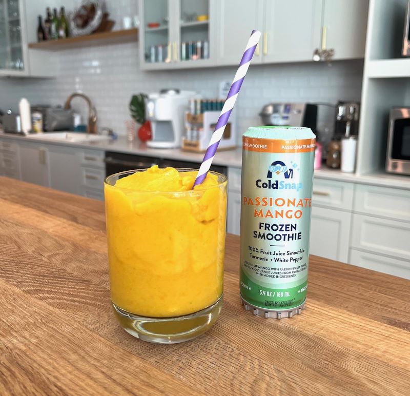 Glass of yellow frozen smoothie with a pod of ColdSnap Passionate Mango Frozen Smoothie on a kitchen counter.
