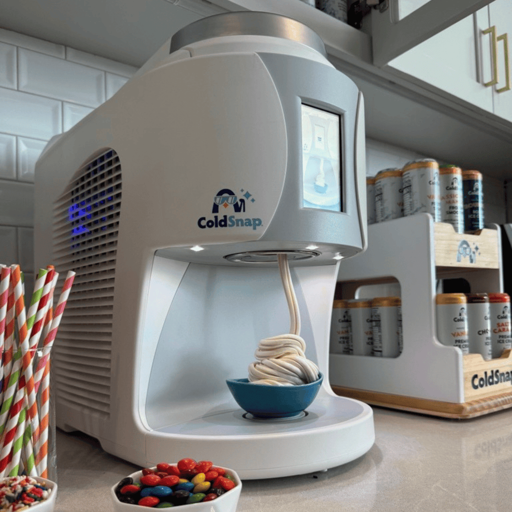 ColdSnap® frozen treat machine sitting on a kitchenette counter next to a ColdSnap® pod display rack and toppings