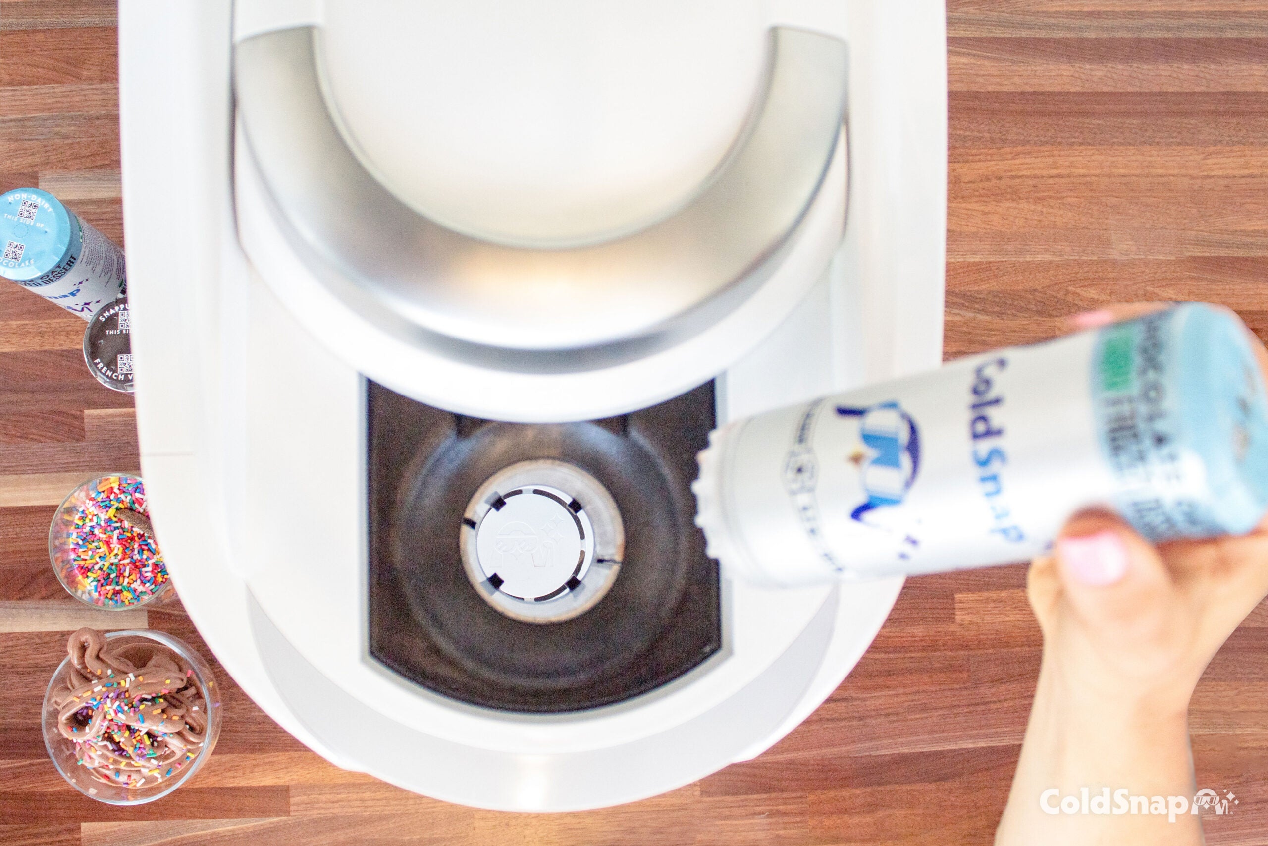 A top view of a ColdSnap ice cream pod being inserted into a ColdSnap machine.  A bowl to the left of the machine that displays a bowl of chocolate ColdSnap ice cream with colorful sprinkles on top. Another bowl above the with colorful sprinkles, and two pods above that. 