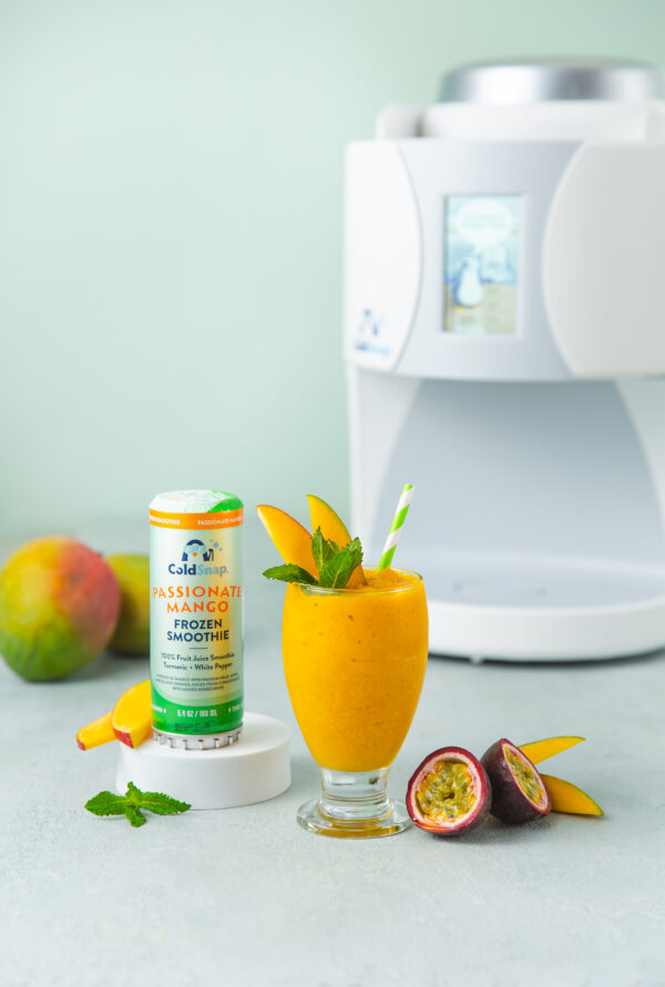 Yellow smoothie with mango, passion fruit, and a ColdSnap Passionate Mano Frozen Smoothie pod on a light background. With a ColdSnap machine in the foreground.