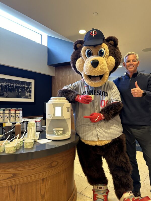 Minnesota Twins Bear Mascot holding a bowl of ColdSnap Ice cream pointing at a ColdSnap machine, with a cheerful man standing next to the mascot with his thumb up.