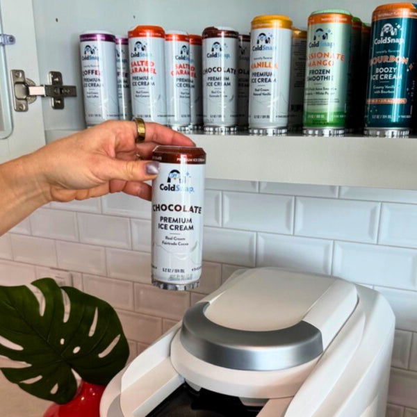 Hand holding a container of ColdSnap Premium Chocolate Ice Cream in front of shelves with assorted ColdSnap ice cream pods.
