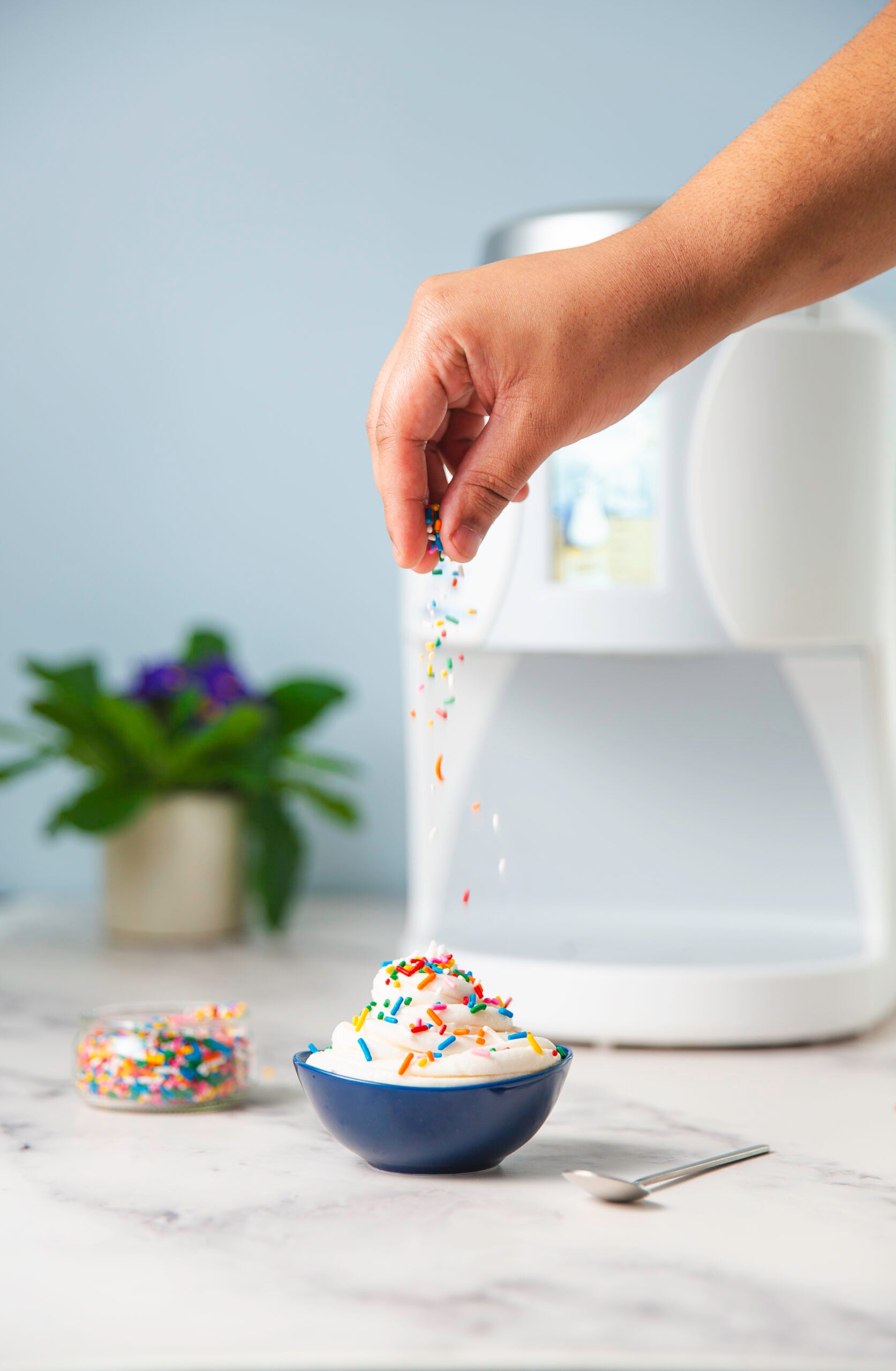 Hand sprinkling colorful sprinkles onto a bowl of ColdSnap premium vanilla ice cream in a blue bowl on a marble surface.