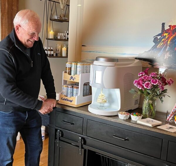 Man standing next to a ColdSnap machine on a kitchen counter with a vase of flowers, bowls of ice cream toppings, and display of assorted ColdSnap pods.